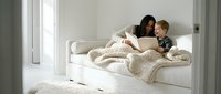 Young Caucasian mother and toddler son reading a book together on a white sofa, wrapped in a cozy knit blanket in a bright living room.