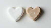 Two heart-shaped natural soap bars in white and beige tones placed side by side on a light background, showcasing organic skincare products.