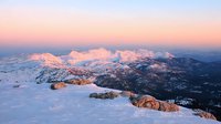 Majestic snow-covered mountain peaks glowing in pink sunset light, aerial winter landscape view of rocky ridges and forested valleys beneath dramatic alpine scenery.