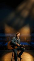 Middle-aged Caucasian male musician with acoustic guitar sitting on stool in empty concert hall under dramatic stage lighting for music performance.
