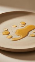 Golden honey drips and puddles on neutral ceramic plate, creating organic flowing patterns. Minimalist food styling with warm tones and soft shadows, suitable for culinary, wellness, and lifestyle design projects.