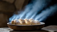 Fresh hot bread rolls steaming in rustic metal pan on wooden table with dramatic lighting and rising vapor creating moody atmospheric food photography.