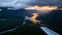Aerial view from airplane window showing dramatic sunset over winding river through forested valley with snow-capped mountains and moody storm clouds.