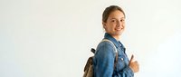 Young woman with brown hair and fair complexion wearing denim shirt and backpack, smiling while giving thumbs up gesture. Student or traveler lifestyle portrait.