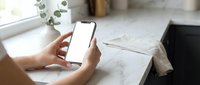 Woman hands holding smartphone with blank white screen mockup on marble table with eucalyptus branch and notebooks in bright home office workspace.