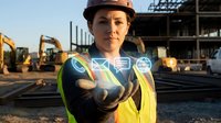 Female construction worker wearing safety helmet and reflective vest pointing at camera on active job site with heavy machinery and steel framework in background.