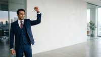 Confident businessman in navy suit celebrating success with fist pump gesture in modern office space with city skyline view, corporate achievement and professional victory concept.