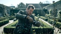 Mature male gardener with tattoos crouching among hedges in formal garden, wearing work clothes, examining plants with focused expression and muddy hands during outdoor landscaping work.