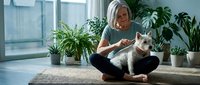 Senior woman with gray hair and light skin tone sitting cross-legged on floor playing with white cat among potted plants by window, indoor pet care and companionship.