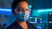 Female scientist wearing protective mask and safety glasses in modern laboratory with blue LED lighting for research and medical testing.