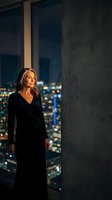 Elegant blonde businesswoman in black dress standing by floor-to-ceiling windows overlooking illuminated city skyline at night in modern office building.