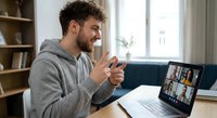 Young bearded man in gray hoodie having video conference call on laptop at home office, remote work meeting with colleagues on screen.