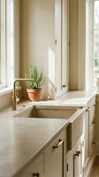 Minimalist kitchen sink area with cream cabinetry, marble countertop, brass faucet, and potted rosemary plant by bright windows with natural light streaming through.