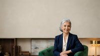 Mature woman with short gray hair and light skin tone, wearing dark blazer and white shirt, sitting on green chair in modern office setting, smiling at camera, professional business portrait for corporate communications and leadership representation.