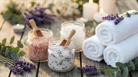 Spa salt scrubs with lavender and eucalyptus herbs, rolled towels, and candles arranged on wooden surface for wellness and relaxation treatments.