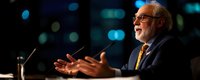 Senior businessman with glasses and beard speaking at conference podium with microphone against blurred city lights background during evening presentation.
