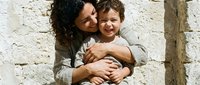 Happy mixed-race mother embracing laughing toddler son against rustic stone wall background. Warm family bonding moment for parenting and lifestyle content.