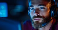 Bearded male gamer in headset illuminated by blue monitor light, focused expression while playing competitive esport game in dark room. Professional streamer setup.