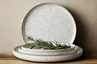 Minimalist ceramic dinnerware stack with speckled white glaze and rosemary sprig on wooden surface against neutral background for kitchen styling.
