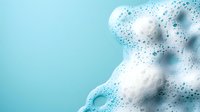 White soap foam with bubbles on turquoise background, macro photography showing texture and pattern of cleaning product or shampoo lather.