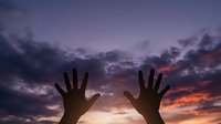 Silhouette of human hands reaching upward against dramatic purple and orange sunset sky with clouds for spiritual and freedom concepts.