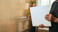 Man holding blank white canvas in warm sunlit room with plants, suitable for mockup, advertising, and creative portfolio presentations.