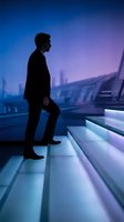 Silhouetted male professional in dark suit climbing illuminated steps with urban skyline in background, representing career growth and business ambition.