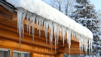 Long icicles hanging from snow-covered wooden cabin roof edge against winter forest background, dramatic natural ice formation creates seasonal atmosphere.