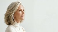 Mature woman with shoulder-length blonde hair wearing cream turtleneck, looking over shoulder against white background for portrait and lifestyle use.