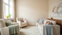 Modern minimalist living room interior with neutral beige walls, comfortable sectional sofa, cozy armchair, soft pastel throws and natural light from windows.