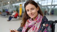 Young Caucasian woman with long brown hair wearing floral pink scarf and navy jacket smiling while using smartphone in airport terminal waiting area.