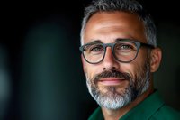 Confident mature Caucasian businessman with gray beard and stylish glasses wearing green blazer smiles warmly against dark background. Professional headshot portrait.