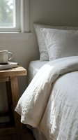 Cozy bedroom corner with white linen bedding, wooden nightstand, metal mug, and natural light from window for home decor and lifestyle design.