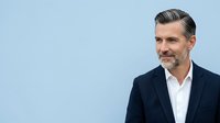 Middle-aged man with gray hair and facial hair wearing navy blazer and white shirt against light blue background for corporate and business use.