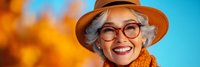 Cheerful senior woman with gray hair wearing stylish hat and glasses smiling warmly against vibrant autumn background for lifestyle content.