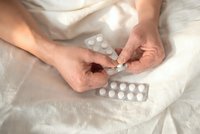 Adult hands holding white medication pills over bed sheets with blister packs nearby for healthcare and treatment concepts.