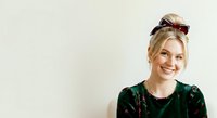 Young woman with blonde hair and fair complexion wearing dark green velvet top with sunglasses on head, smiling against neutral background for portrait and lifestyle use.