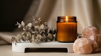 Spa wellness setup with burning candle in amber glass jar, himalayan salt stones and eucalyptus branch creating relaxing aromatherapy atmosphere.