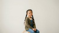 Young girl with dark braided hair and mixed ethnicity, wearing olive green cardigan over denim overalls, sitting against neutral background for portrait and lifestyle use.