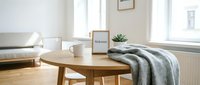 Cozy minimalist living room with wooden round table, welcome sign, mug, gray blanket, and succulent plant by bright window.