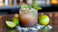 Fresh mojito cocktail with crushed ice, whole limes, mint leaves and garnish in textured glass on dark wooden surface against blurred bar background.