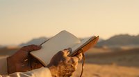Young adult male with dark skin tone holding tablet device and stylus in arid landscape at golden hour, suitable for technology, education, and outdoor professional concepts.