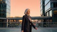 Senior woman with gray hair wearing dark blazer standing in modern urban plaza at golden hour sunset, contemporary office buildings surrounding, professional business district setting.
