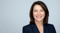 Professional middle-aged Caucasian businesswoman in navy blazer smiling confidently against gray background for corporate headshots and marketing materials.