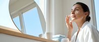 Young woman with South Asian ethnicity applying skincare moisturizer or cosmetic product to face near bathroom mirror and window. Wellness and beauty routine.