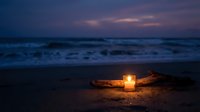 Glowing candle on sandy beach at twilight with ocean waves and purple sunset sky creating peaceful romantic atmosphere for meditation and relaxation.