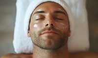 Caucasian man with stubble relaxing during spa facial treatment, wearing white towel on head, eyes closed with moisturizing cream on cheeks.