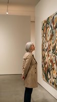 Senior woman with gray hair in beige coat admiring colorful abstract artwork in modern art gallery museum exhibition space.