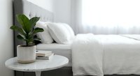 Modern minimalist bedroom interior with white bedding, marble nightstand, green plant in ceramic pot, and natural light from window creating serene atmosphere.