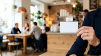 Person in dark business attire holding blank white card in modern cafe with wooden tables, plants, and blurred colleagues in background, suitable for corporate networking and business communication.